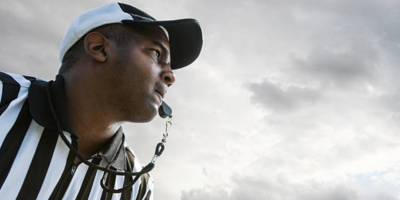 View from below of referee in striped black and white uniform and hat with whistle in his mouth against a clouded sky